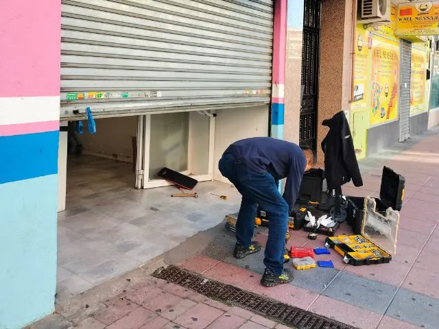 Cerrajero reparando cierre metálico enrollable de comercio, con herramientas profesionales en la vía pública.